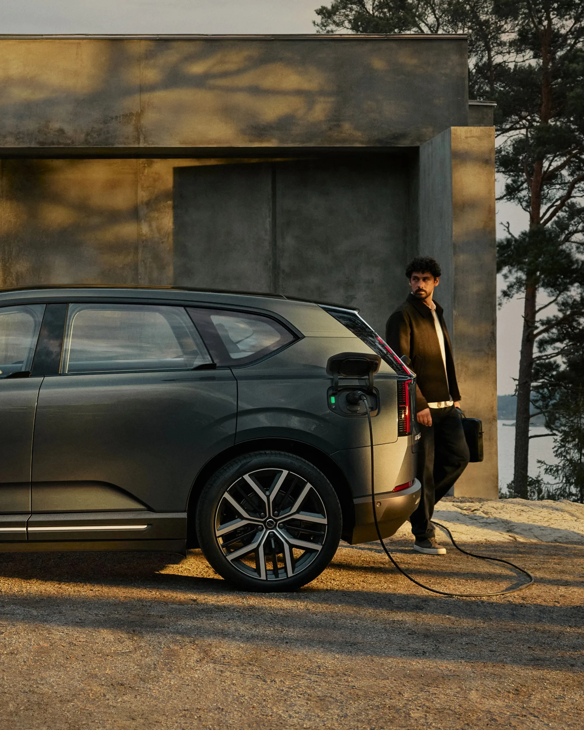 A person stands beside a Volvo EX60 plugged into a home charger at dusk.